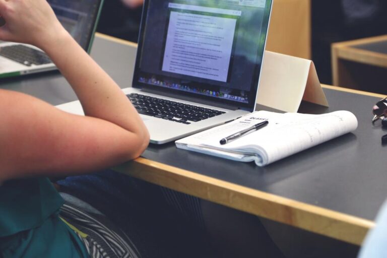 A high school student reviewing work on a laptop with a notebook and pen on the desk.