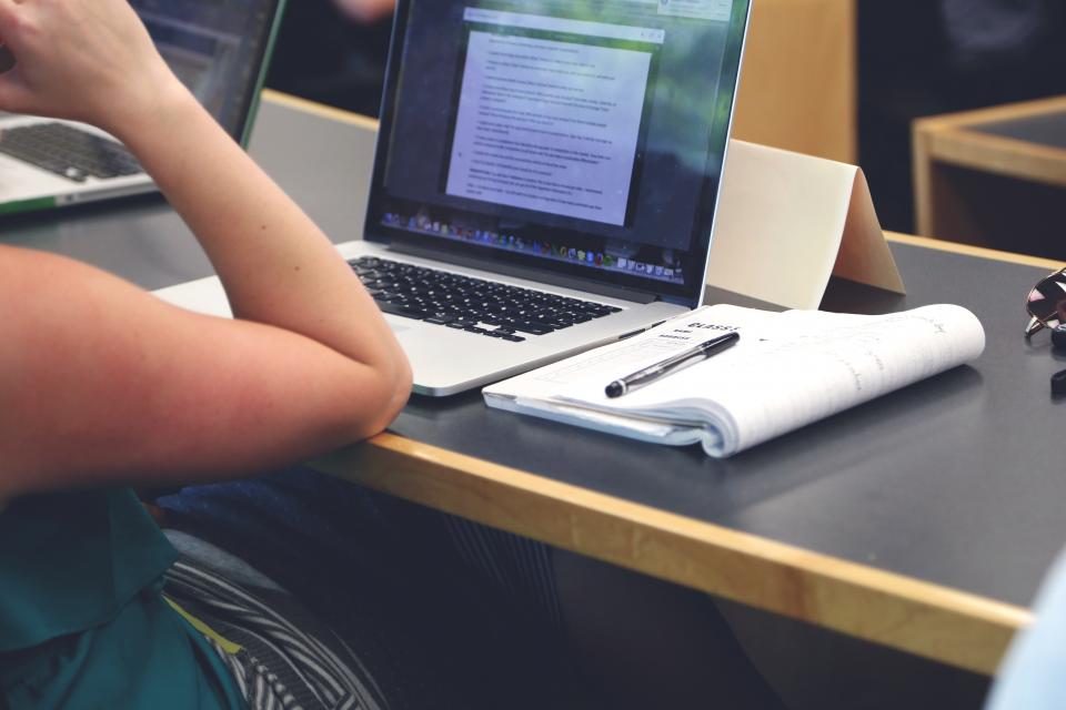 A high school student reviewing work on a laptop with a notebook and pen on the desk.